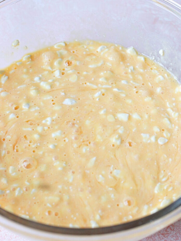 A clear glass bowl filled with a thick, creamy, pale orange batter that contains visible small white lumps, suggesting it may include cottage cheese or curdled ingredients.