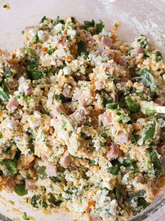 A close-up of a glass bowl filled with a chunky mixture of eggs, chopped ham, spinach, shredded cheese, and herbs, ready to be cooked or baked.