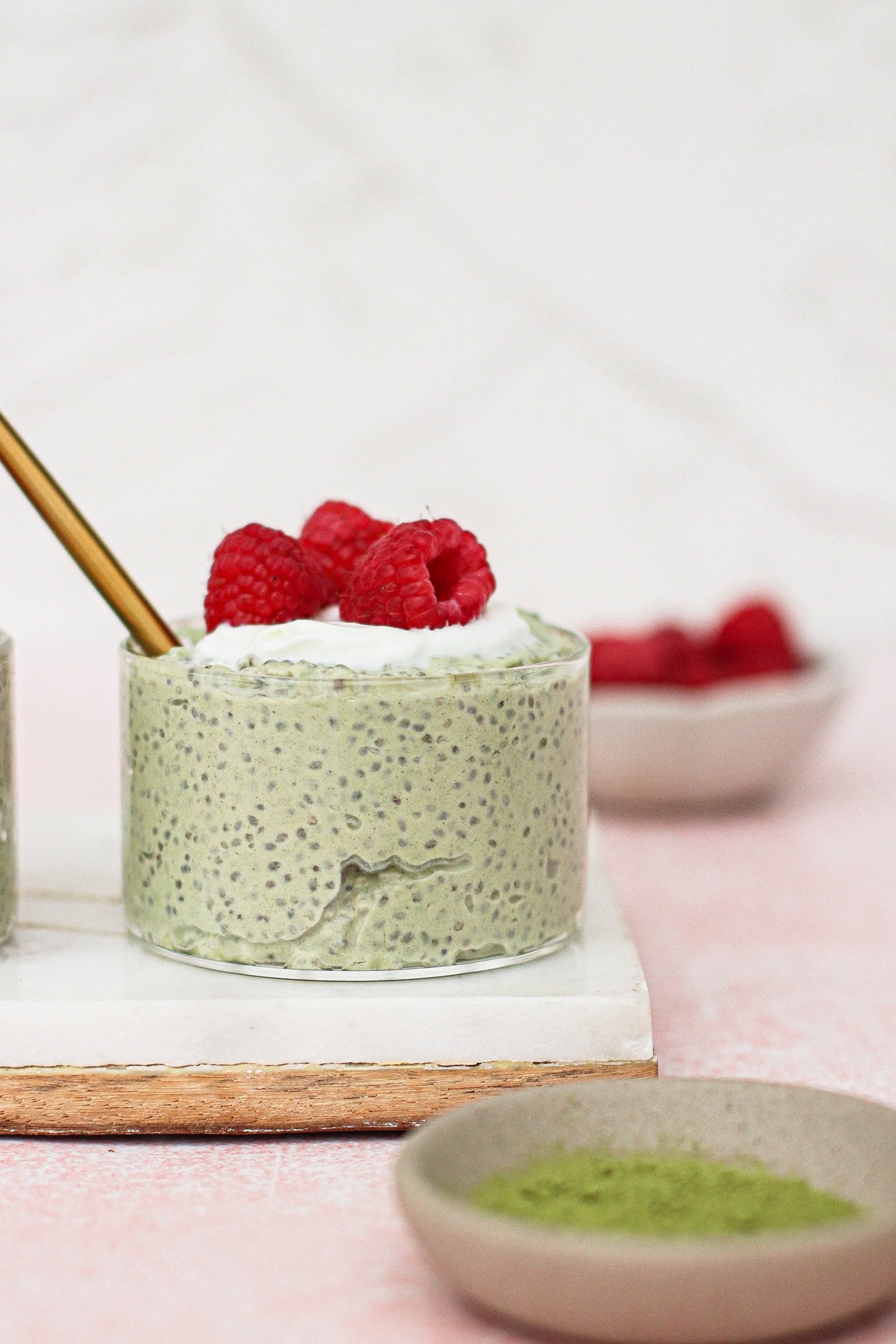 A small round dish of matcha chia pudding topped with whipped cream and fresh raspberries sits on a white board. A gold spoon is in the pudding, and bowls of raspberries and green powder are nearby.