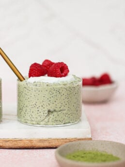 A small glass of matcha chia pudding topped with whipped cream and fresh raspberries sits on a white tray. A gold spoon is inside the glass. Bowls with raspberries and green powder are nearby against a light pink background.