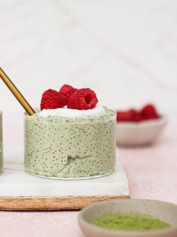 A small glass of matcha chia pudding topped with whipped cream and fresh raspberries sits on a white tray. A gold spoon is inside the glass. Bowls with raspberries and green powder are nearby against a light pink background.