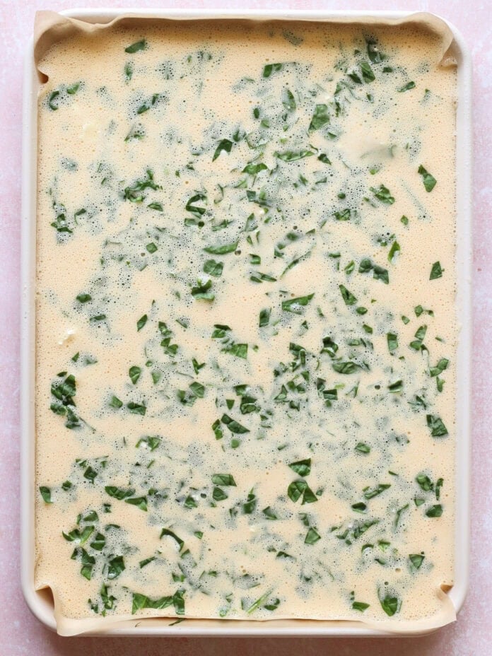 A baking sheet lined with parchment paper, filled with an uncooked egg mixture and chopped green herbs, ready to be baked.