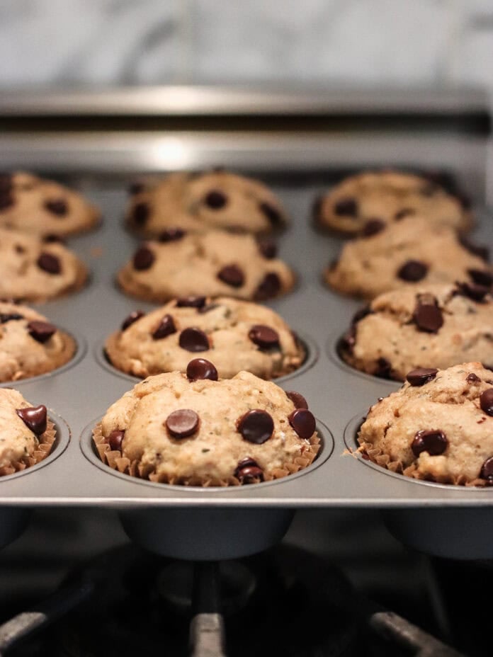 A muffin pan filled with freshly baked chocolate chip muffins sits on a stovetop. The muffins are golden brown and studded with melted chocolate chips.