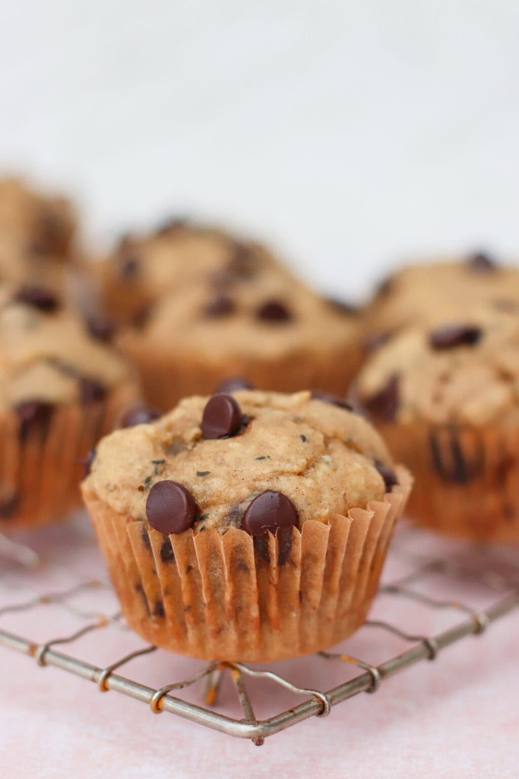 A close-up of chocolate chip muffins in brown paper liners, resting on a cooling rack. The muffins are golden brown with chocolate chips on top, and more muffins are blurred in the background.