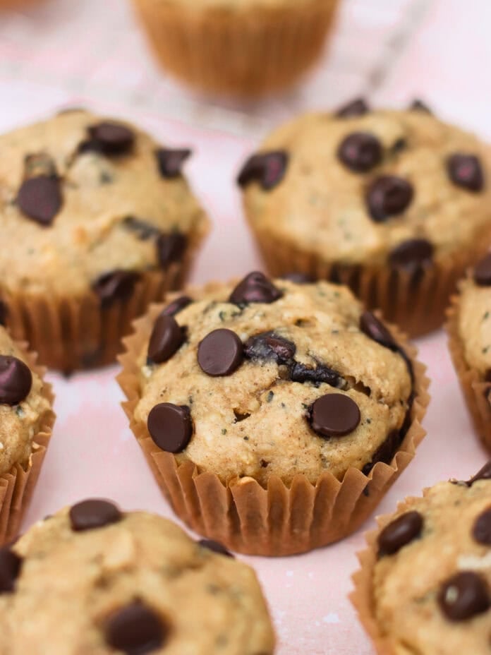 Close-up of several banana protein muffins with chocolate chips in brown paper liners, arranged on a light pink surface. The muffins are golden brown with visible chocolate chips on top.