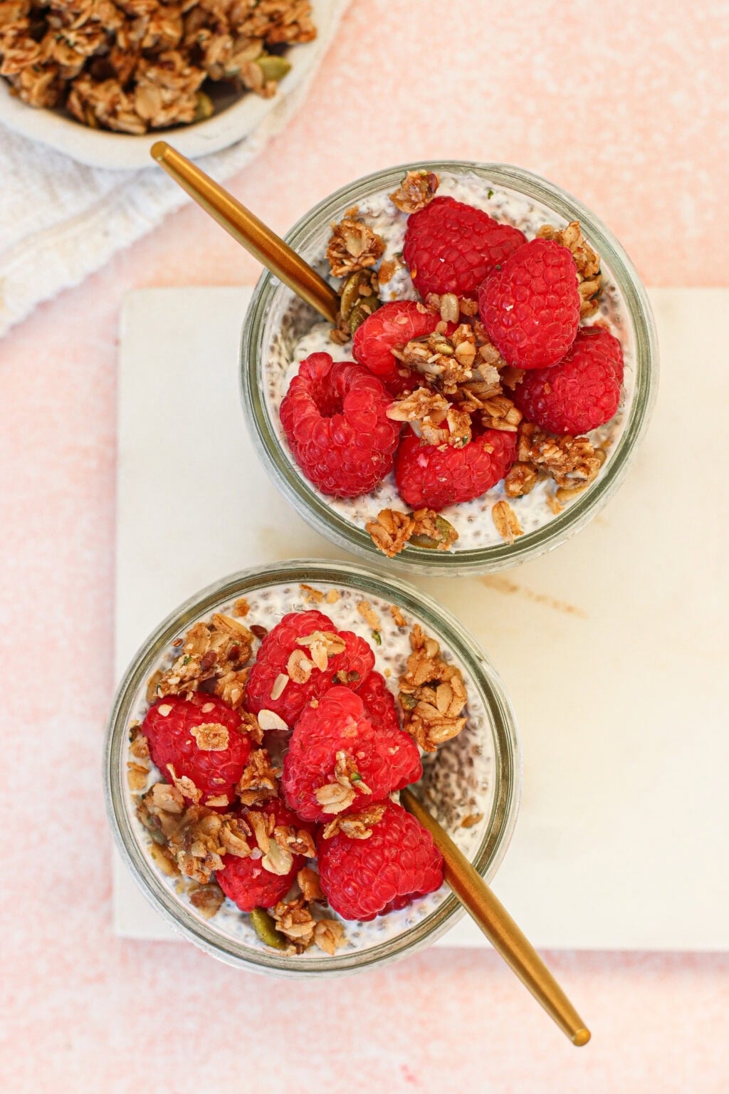 Two glass jars filled with chia pudding, topped with fresh raspberries and granola, each with a gold spoon, placed on a light surface with a bowl of granola in the background.