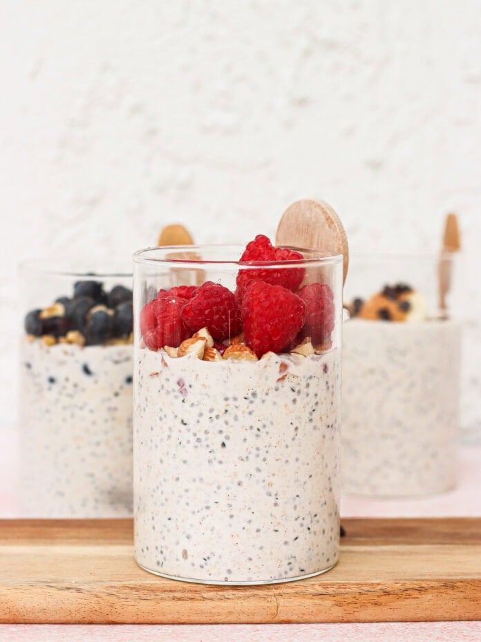 Three glass cups of overnight oats topped with fresh berries and granola sit on a wooden board. The front cup has raspberries, while the back cups have blueberries and wooden spoons in each.