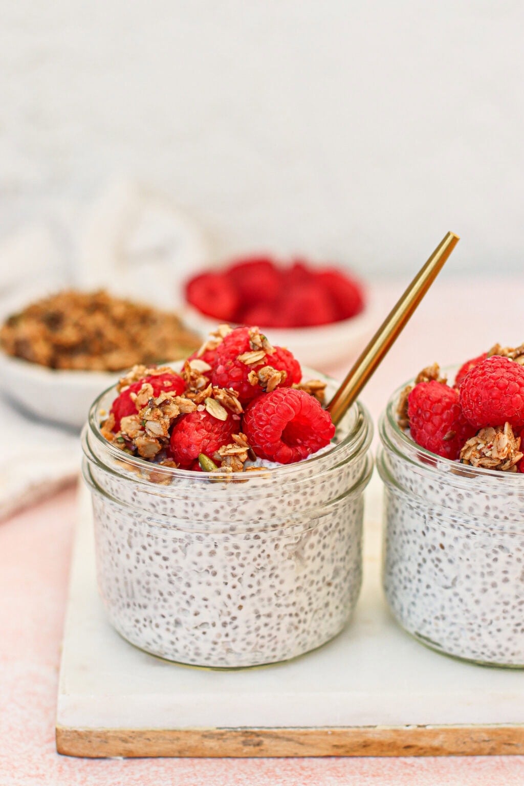 Two glass jars filled with chia pudding are topped with granola and fresh raspberries. A gold spoon rests in one jar. In the background, bowls of granola and raspberries are slightly out of focus.
