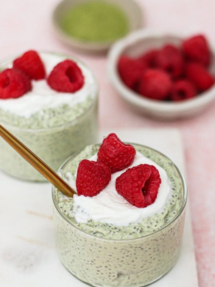 Two glass cups filled with green chia pudding, topped with a dollop of whipped cream and fresh raspberries. A gold spoon rests in one cup. In the background, out-of-focus bowls contain raspberries and green powder.