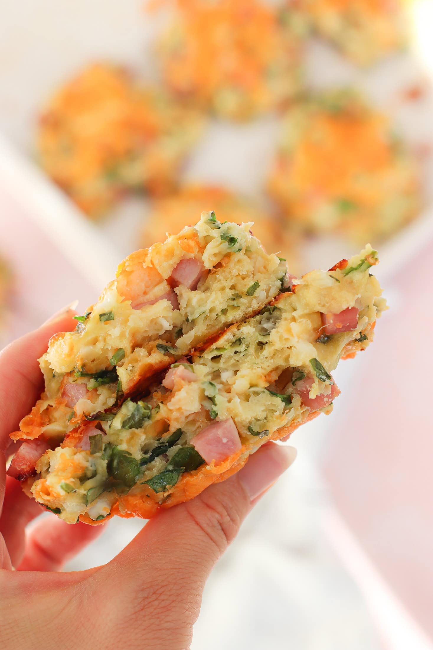 A hand holds a halved savory muffin or fritter, showing a filling of chopped vegetables, herbs, and diced ham. The texture appears fluffy and moist, with visible green and orange ingredients.