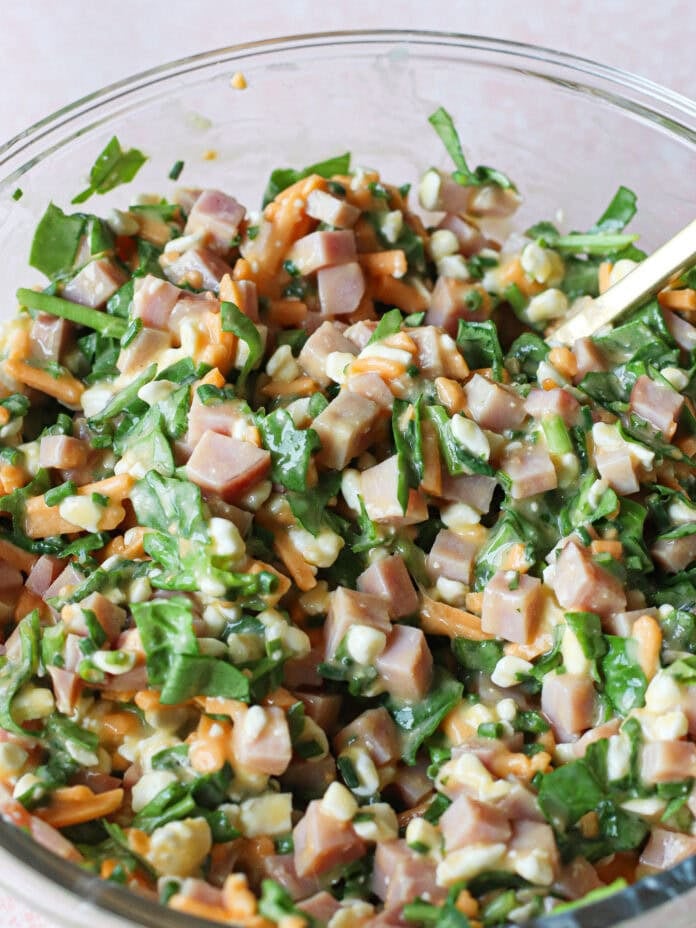 A clear glass bowl filled with a chopped salad containing diced ham, shredded cheese, spinach, and cottage cheese, with a gold spoon partially visible on the side.