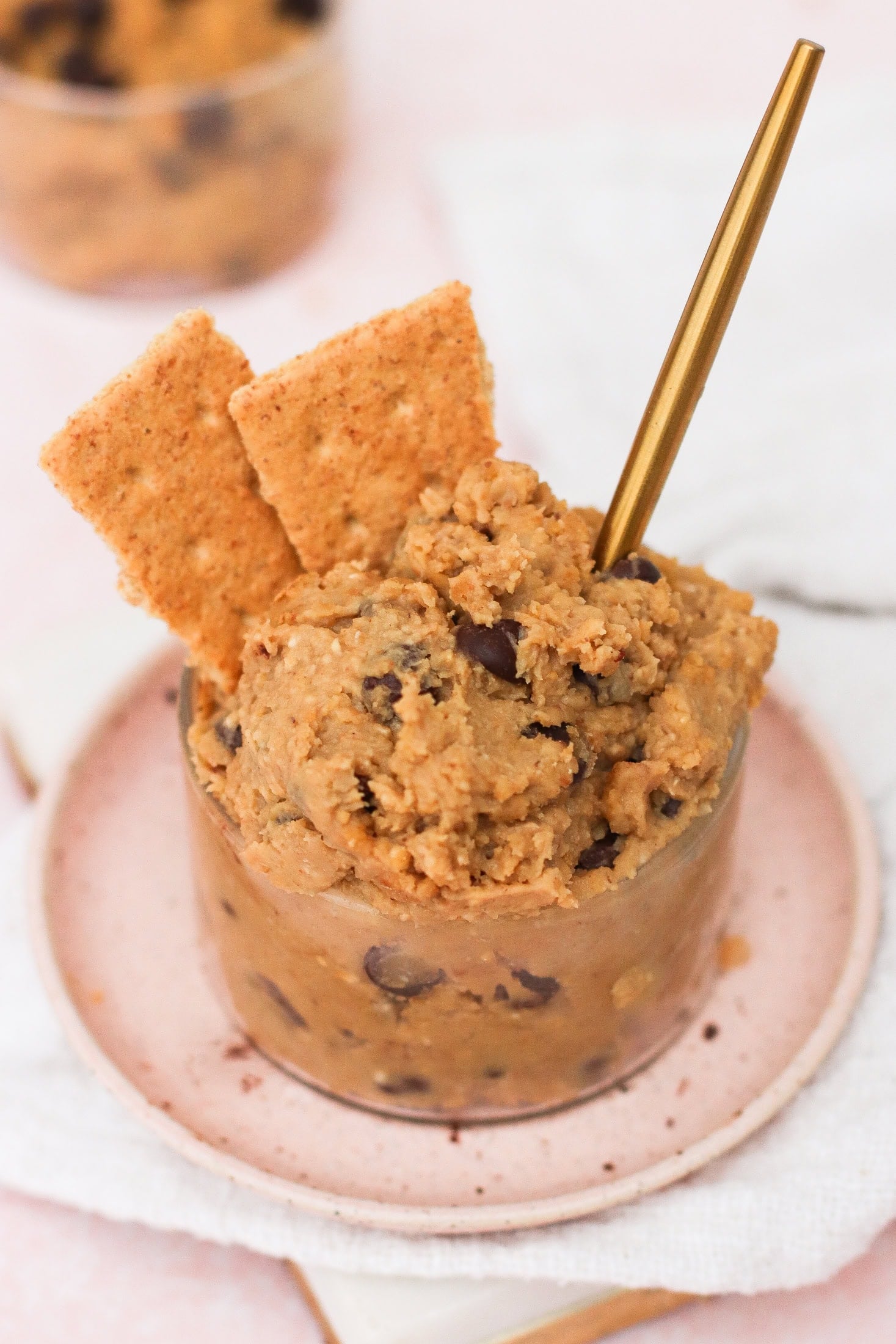 A glass cup filled with chocolate chip cookie dough, garnished with two rectangular graham crackers and a gold spoon, sits on a small pink plate atop a white napkin.