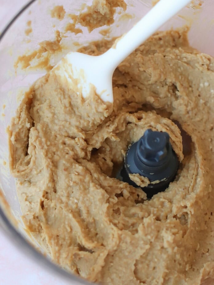 A close-up of thick, beige cookie dough being mixed in a food processor with a white spatula.