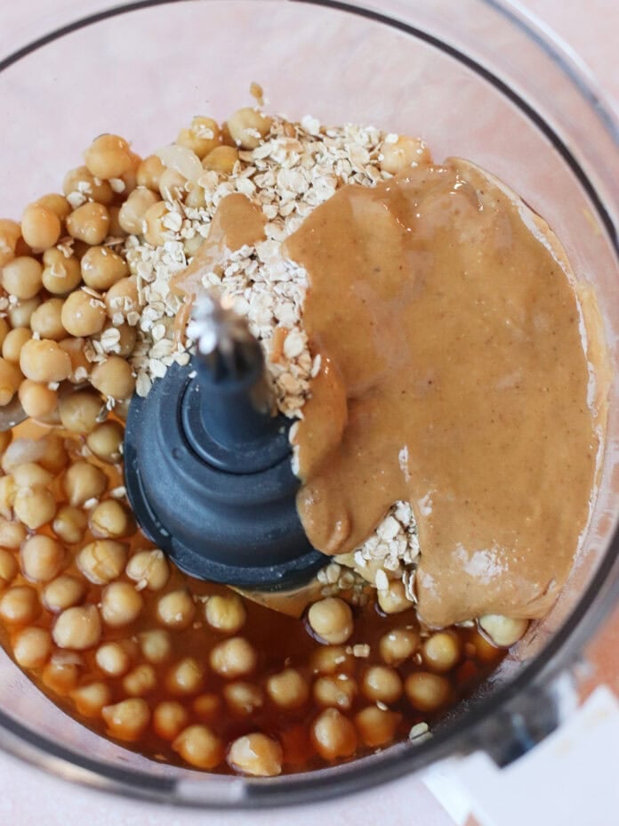 A food processor containing chickpeas, rolled oats, almond butter, and a brown liquid ingredient, viewed from above before blending.