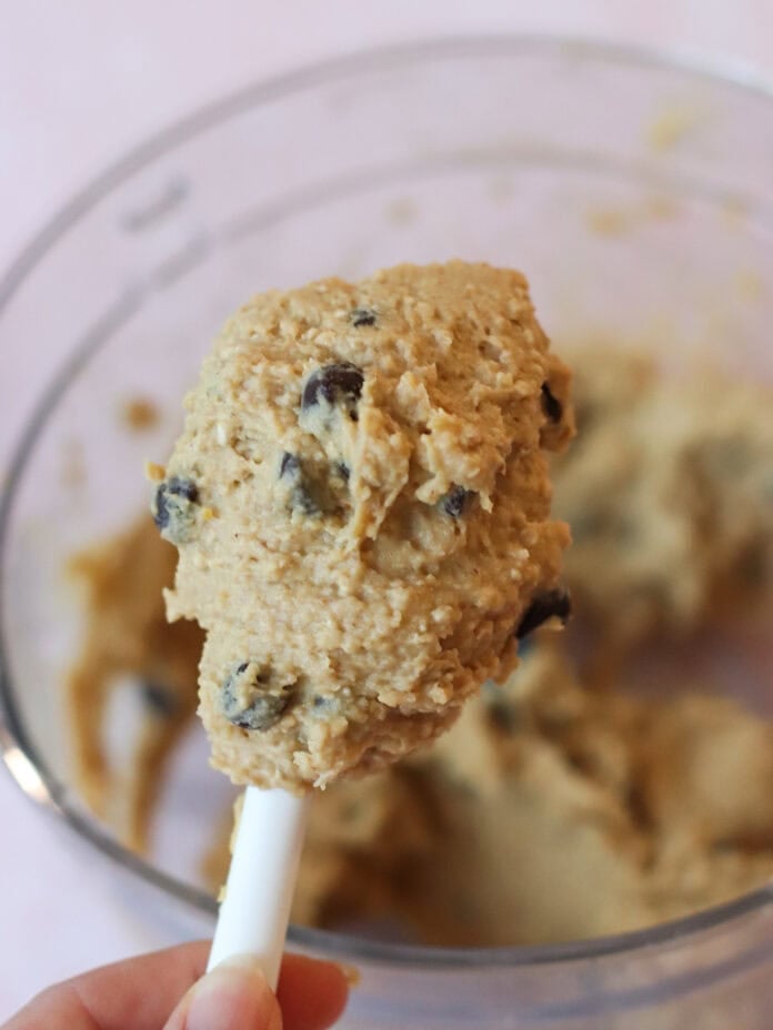 A close-up of a hand holding a spatula with chocolate chip cookie dough above a glass mixing bowl filled with more dough.