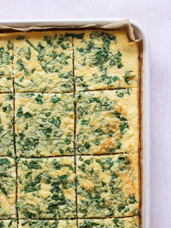 A close-up of a rectangular baking dish filled with baked egg squares topped with chopped green herbs, cut into evenly sized pieces on parchment paper against a pale pink background.