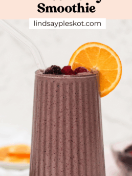 A tall glass of mixed berry smoothie garnished with an orange slice and berries, topped with a glass straw. Text above the smoothie reads: “Healthy High-Fiber Mixed Berry Smoothie; lindsaypleskot.com.”.