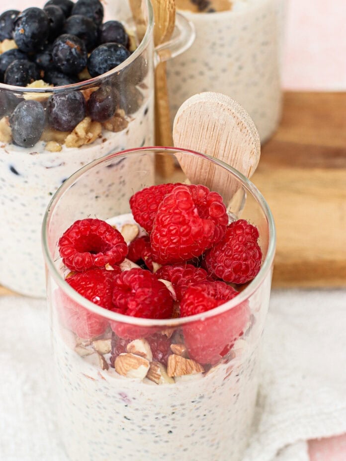 Glass cups filled with overnight oats topped with raspberries, blueberries, and chopped nuts, each with a small wooden spoon. The cups are placed on a white cloth and wooden board.