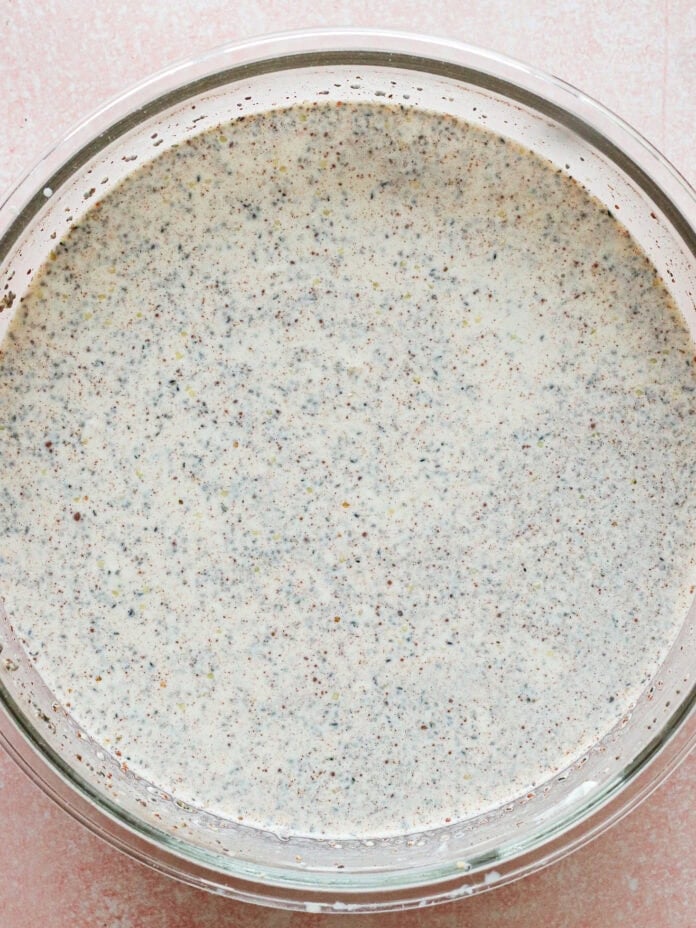 A glass bowl filled with a creamy mixture speckled with small black seeds, likely poppy seeds, sits on a light pink textured surface.