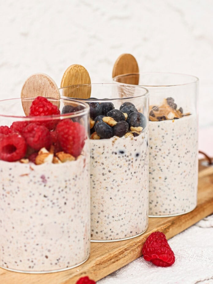 Three glasses of chia seed pudding topped with fresh raspberries, blueberries, and chopped nuts, each with a wooden spoon, displayed on a wooden board with scattered raspberries nearby.