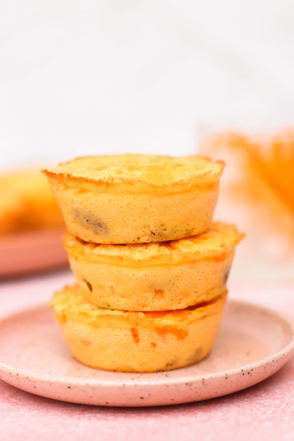 Three mini quiches are stacked on a pink ceramic plate. The quiches have a golden-brown top and bits of filling visible inside the crust. The background is softly blurred with warm tones.