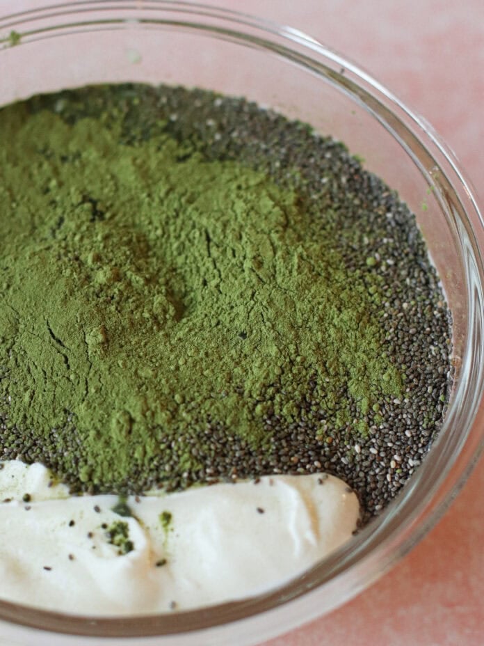 A glass bowl containing chia seeds, green matcha powder, and a dollop of yogurt on a pink surface.