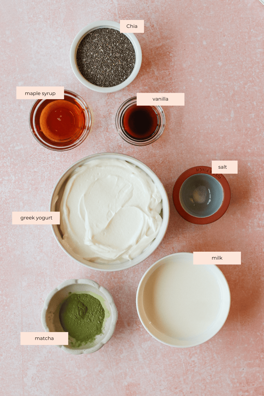 A flat lay of labeled ingredients for matcha chia pudding on a pink surface: chia seeds, maple syrup, vanilla, salt, Greek yogurt, milk, and matcha powder, each in separate bowls.