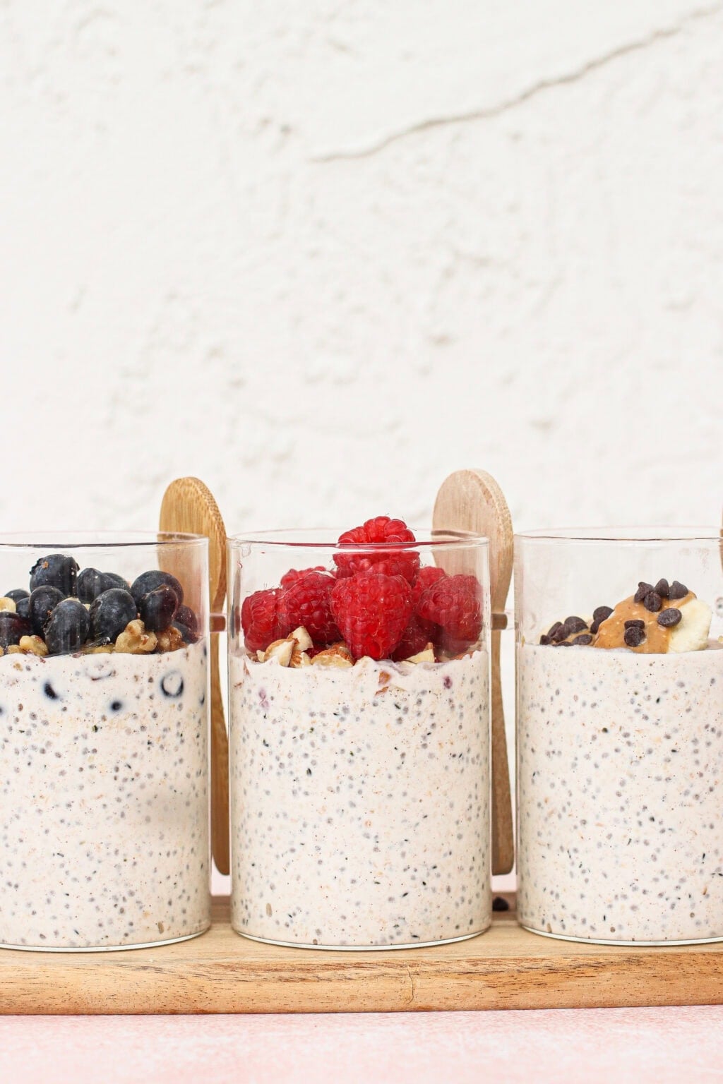 Three clear glasses filled with overnight oats are topped with blueberries, raspberries, and banana slices with chocolate chips. Wooden spoons are placed behind each glass, and the background is a light textured wall.