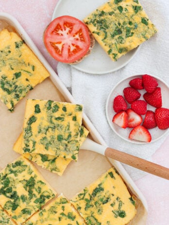A baking dish with sliced spinach frittata, a plate with a frittata square and half a tomato, and a small bowl of raspberries and sliced strawberries on a light pink surface.