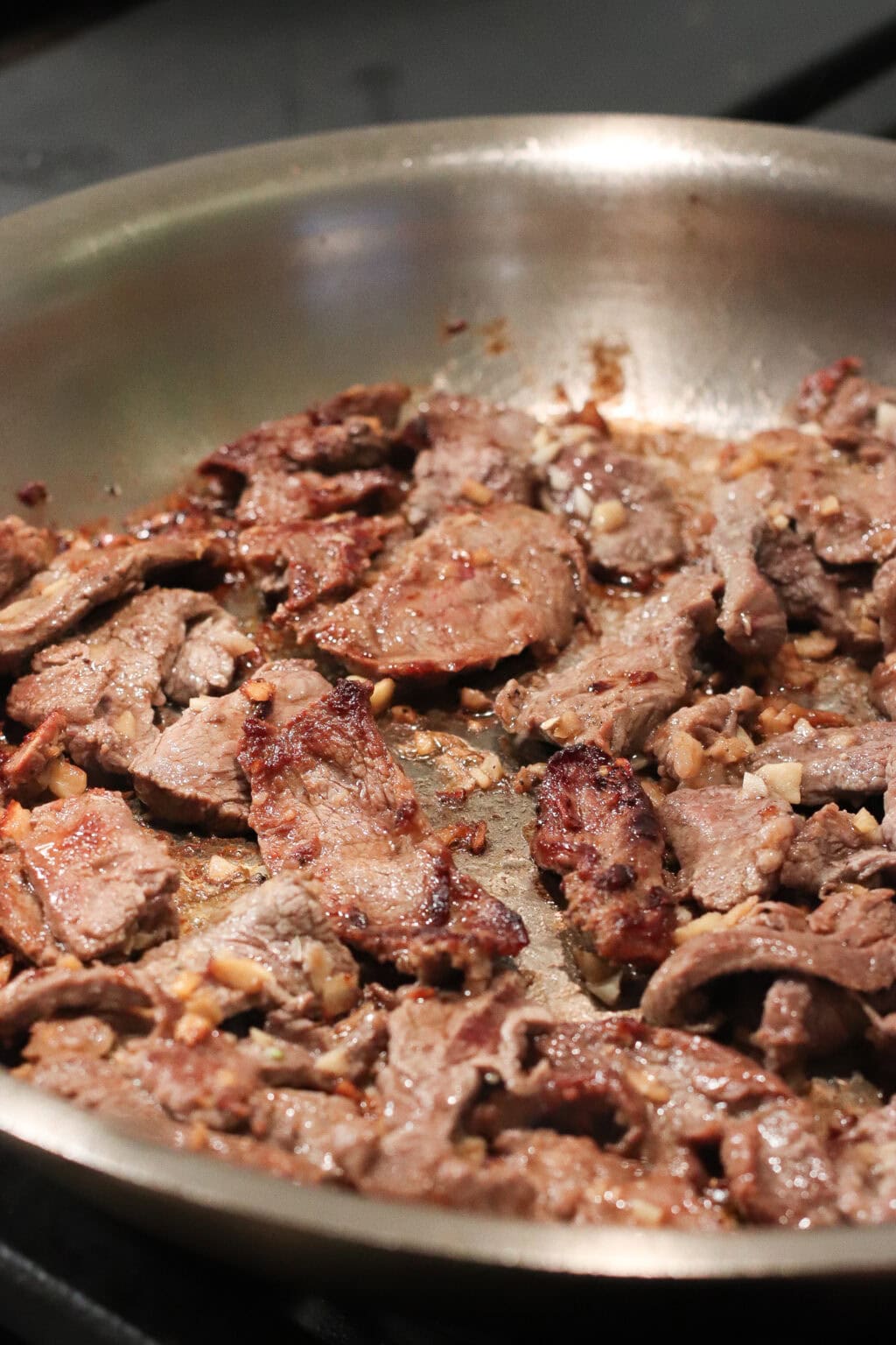 Slices of beef cooking and browning in a stainless steel skillet, with visible pieces of garlic and a caramelized surface on the meat.