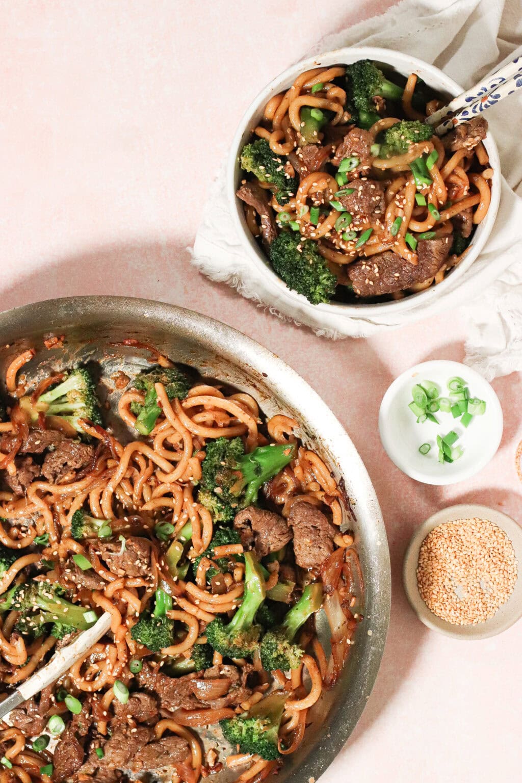 A skillet with beef, broccoli, and noodles in a savory sauce sits beside a bowl of the same dish, garnished with green onions and sesame seeds. Small bowls of green onions and sesame seeds are nearby on a pink surface.