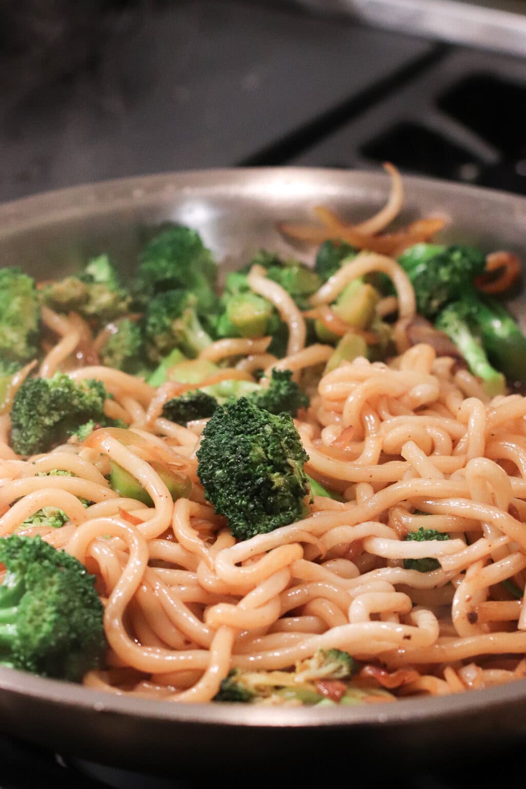 A close-up of a pan filled with stir-fried noodles, broccoli florets, and vegetables, all lightly coated in sauce. Steam rises from the hot dish, indicating it has just been cooked.