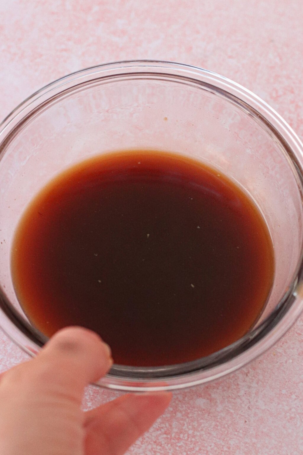 A hand holds a clear glass bowl filled with a dark brown liquid, set on a light pink surface.