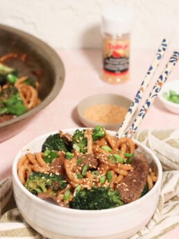 A bowl of stir-fried noodles with beef, broccoli, and onions, topped with sesame seeds and green onions. Chopsticks rest on the bowl, with a skillet, sesame seeds, and seasoning visible in the background.