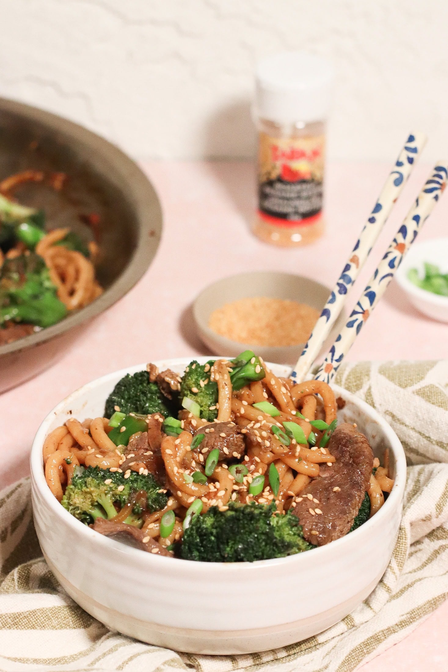 A bowl of stir-fried noodles with beef, broccoli, and onions, topped with sesame seeds and green onions. Chopsticks rest on the bowl, with a skillet, sesame seeds, and seasoning visible in the background.