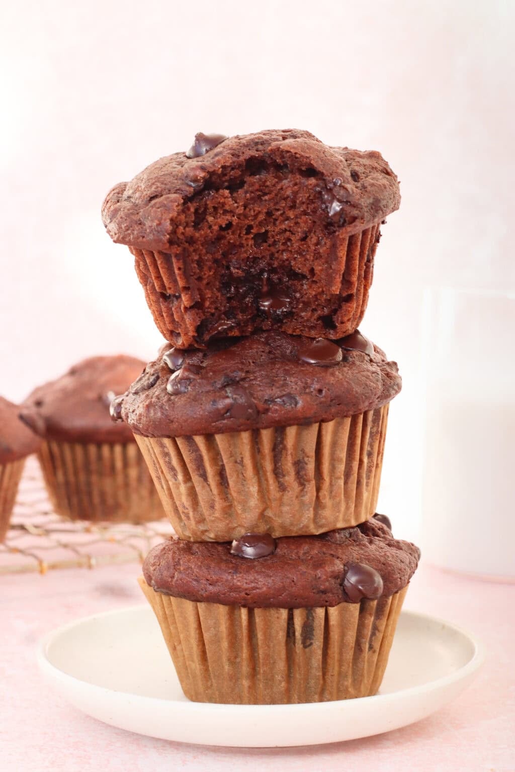 Three chocolate chip muffins are stacked on a white plate, with the top muffin showing a bite taken out. More muffins and a glass of milk are in the background, all set on a light pink surface.