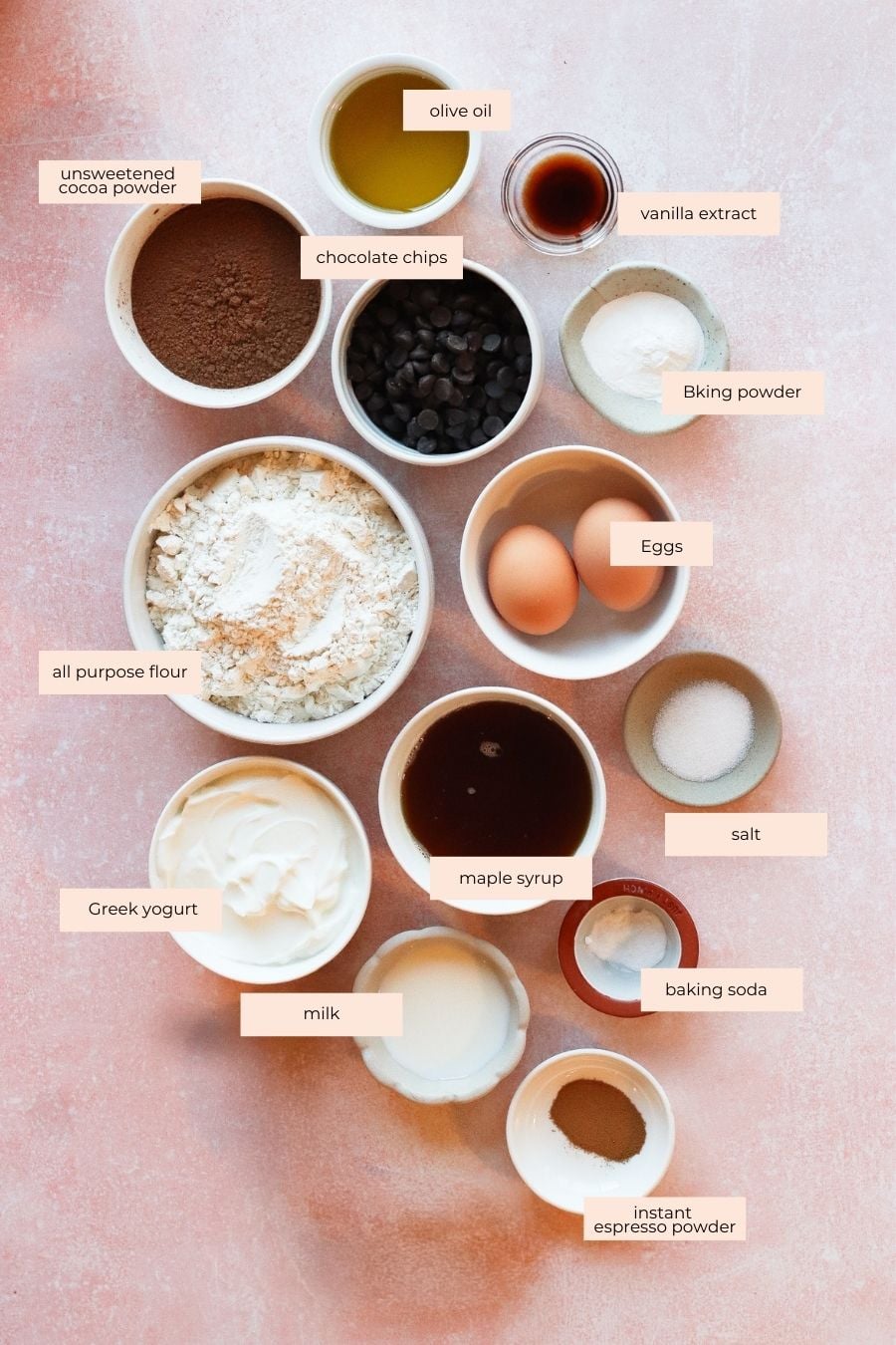 Various baking ingredients for chocolate protein muffins arranged in bowls on a pink surface, including flour, Greek yogurt, eggs, chocolate chips, cocoa powder, maple syrup, milk, olive oil, vanilla extract, baking powder and instant espresso powder.