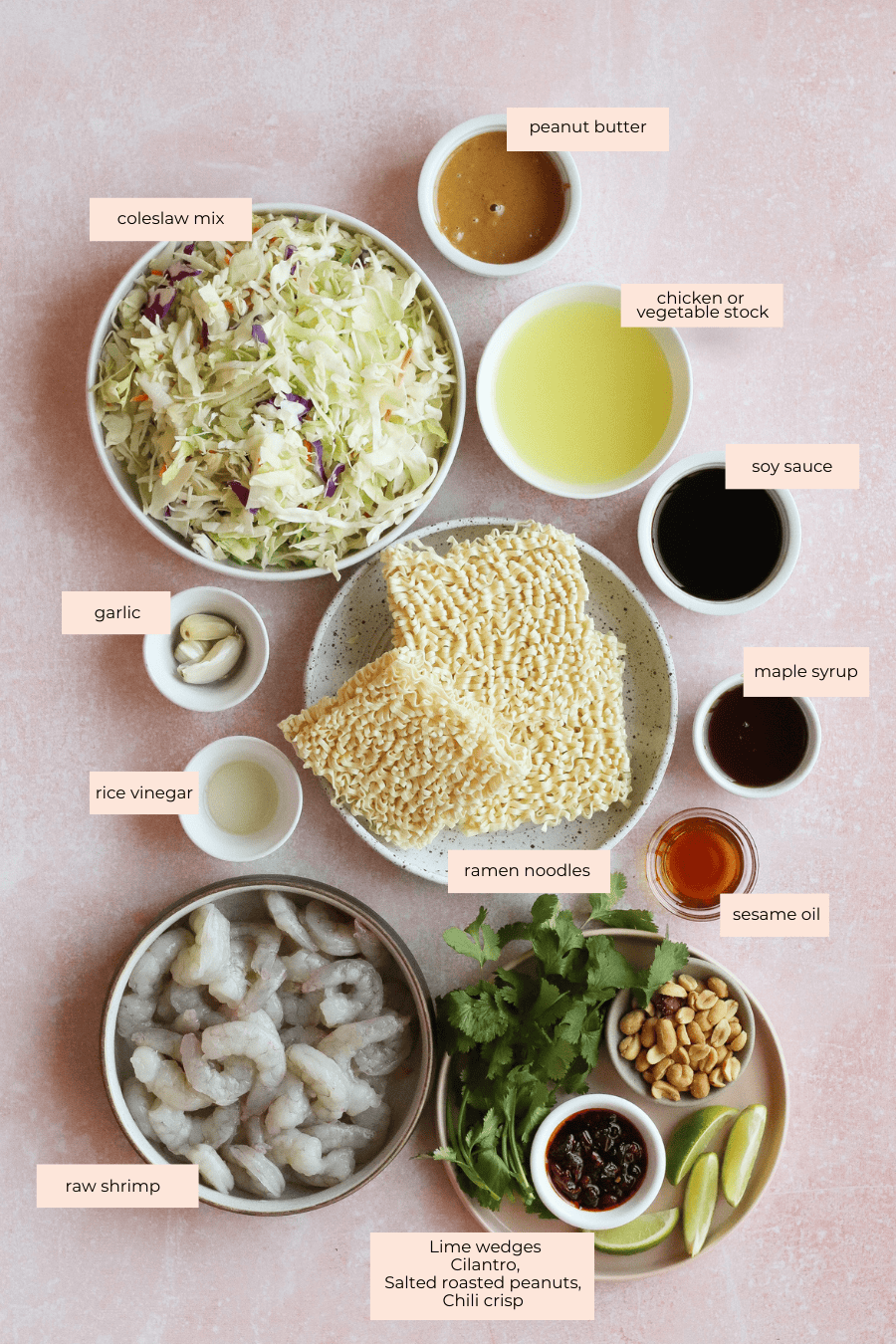 Flat lay of ingredients for a shrimp ramen salad, including coleslaw mix, garlic, rice vinegar, raw shrimp, ramen noodles, peanut butter, chicken stock, soy sauce, maple syrup, sesame oil, cilantro, lime wedges, peanuts, and chili crisp.
