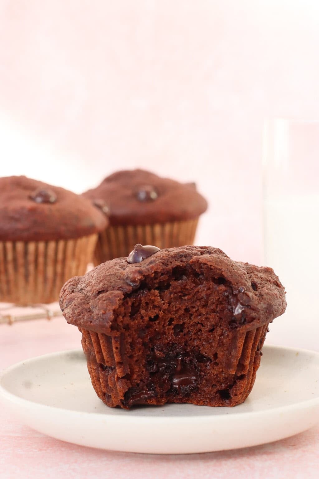 A chocolate muffin with a bite taken out sits on a white plate, revealing a moist, chocolatey inside. Two whole muffins and a glass of milk are in the background on a light pink surface.