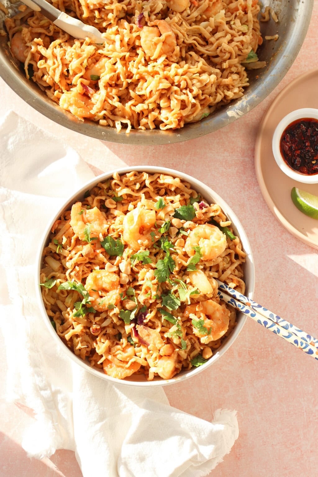 A bowl of shrimp stir-fried noodles garnished with fresh herbs sits on a light surface with a blue-patterned spoon. A larger serving pan and a small dish of sauce with lime are nearby.