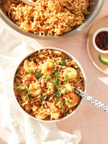 A bowl of shrimp stir-fried noodles garnished with fresh herbs sits on a light surface with a blue-patterned spoon. A larger serving pan and a small dish of sauce with lime are nearby.