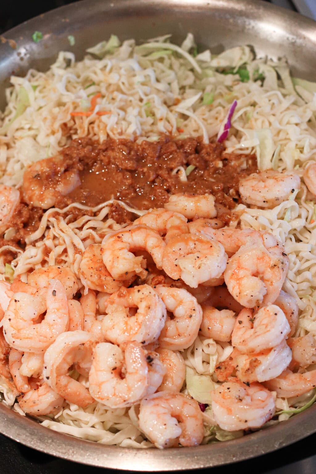 A skillet filled with uncooked shrimp, shredded cabbage, noodles, and a brown sauce, ready to be cooked for a stir-fry dish.
