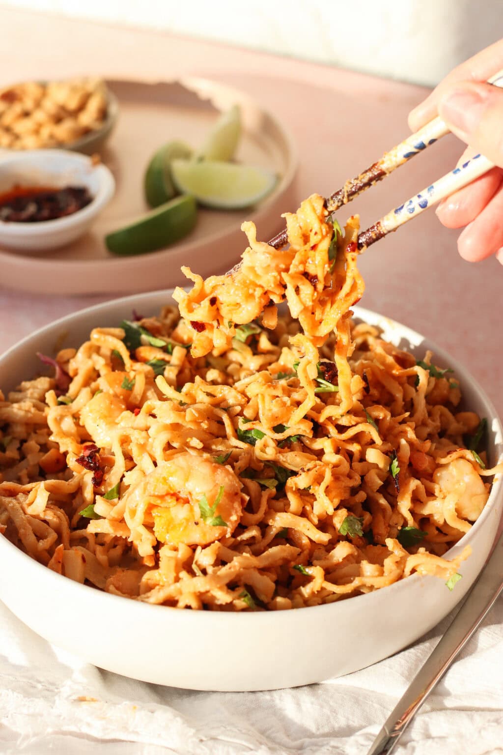 A hand uses chopsticks to lift a portion of noodle salad topped with shrimp and herbs from a bowl. In the background, lime wedges and dipping sauces are visible on a small plate.