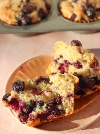 A blueberry muffin cut in half on a plate, showing a moist, fluffy texture with whole blueberries inside. In the background, more muffins are visible in a muffin pan.