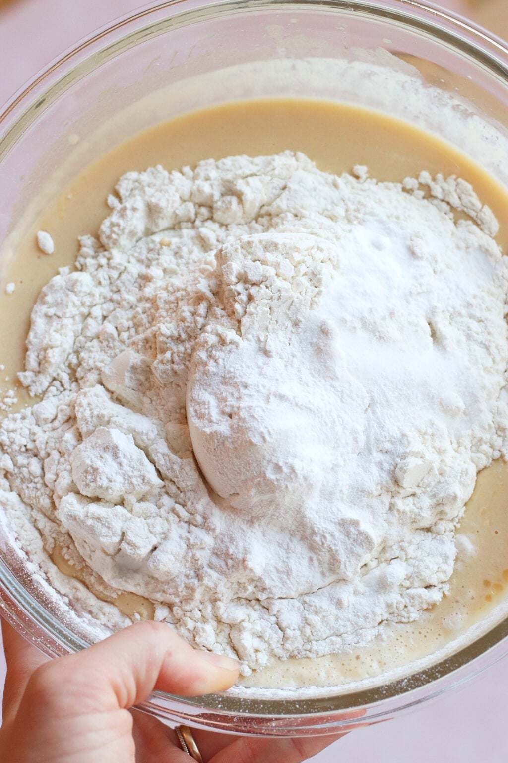 A close-up of a glass bowl containing a creamy batter topped with a mound of flour, held by a hand ready to mix the ingredients together.