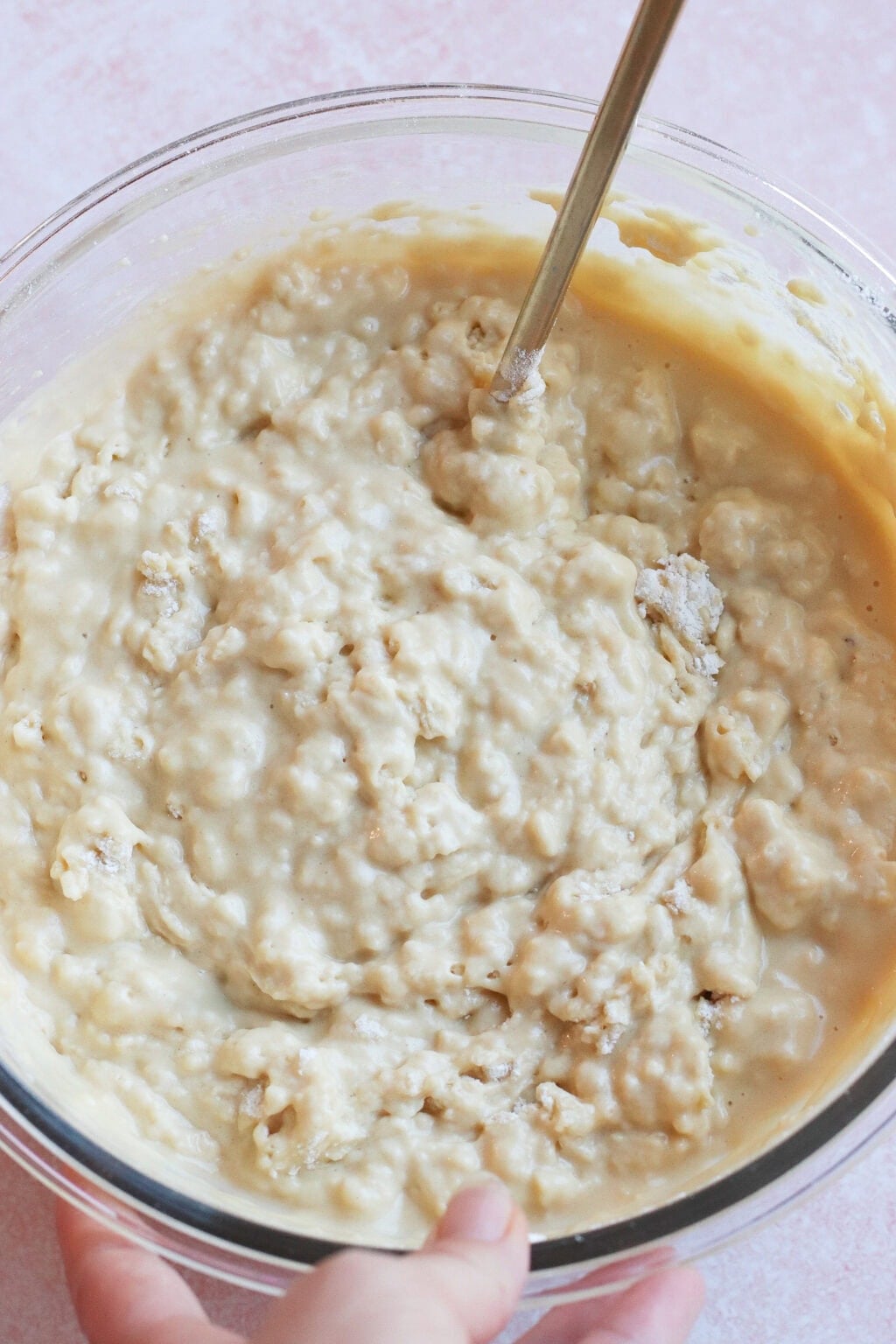 A close-up of a glass bowl filled with a lumpy, pale batter being mixed with a metal spoon. A hand is holding the side of the bowl, and the background is a light pink surface.