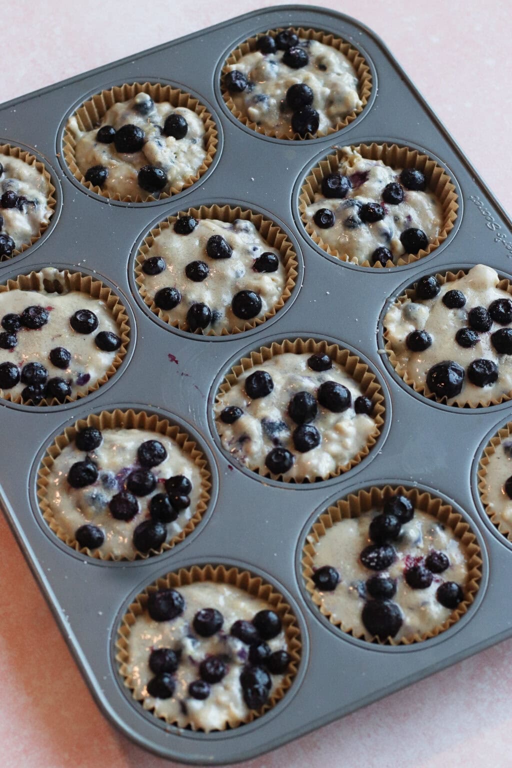 A muffin tin filled with paper liners, each holding unbaked blueberry muffin batter topped with fresh blueberries, ready to be baked.
