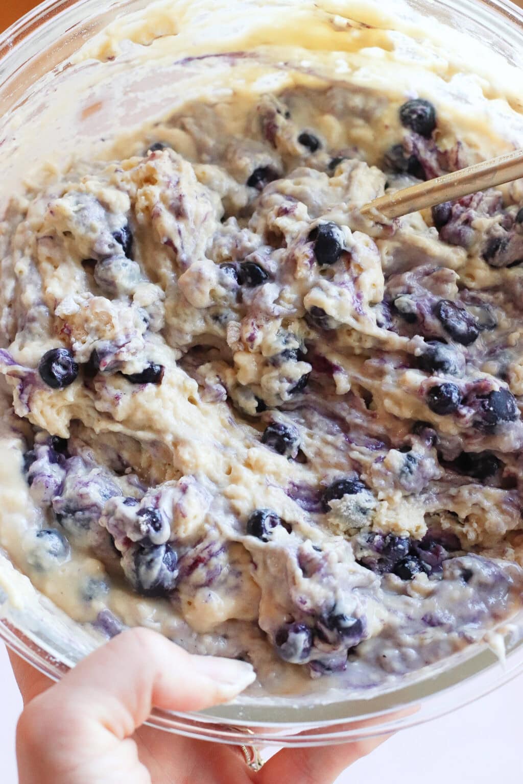 A close-up of a mixing bowl filled with thick blueberry muffin batter being stirred with a spoon, with visible blueberries throughout the mixture.