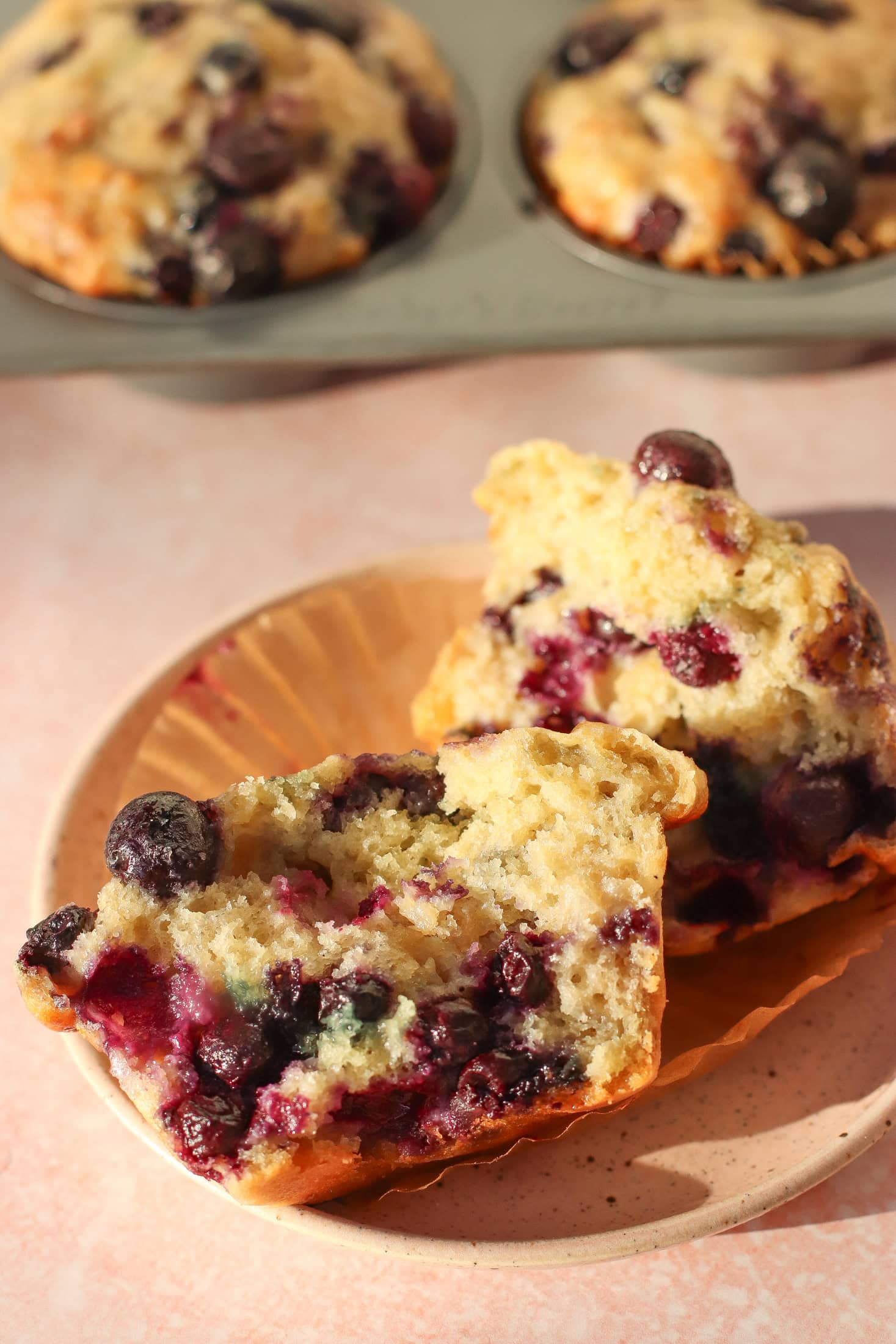 A blueberry muffin cut in half on a plate, showing a moist, fluffy texture with whole blueberries inside. In the background, more muffins are visible in a muffin pan.