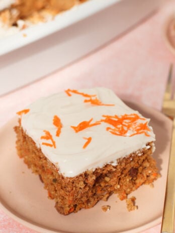 A slice of carrot cake with white frosting and shredded carrots on top sits on a pink plate beside a gold fork.